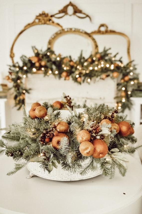 Decorative centerpiece of evergreen branches, oranges, and berries on a table, with a gold-framed mirror and garland in the background.