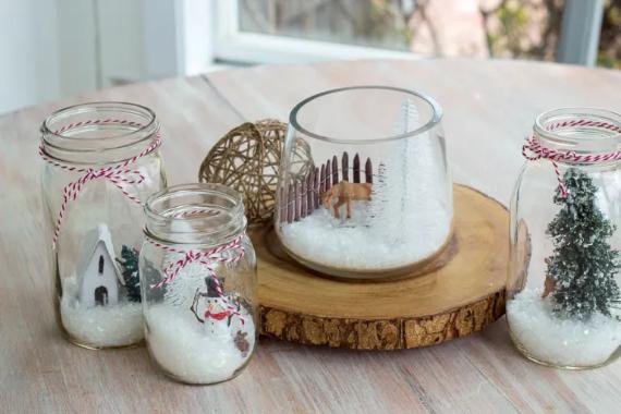 Glass jars on a wooden slab contain snowy winter scenes: a house, snowman, and trees. A cozy decorative setting with a festive feel.