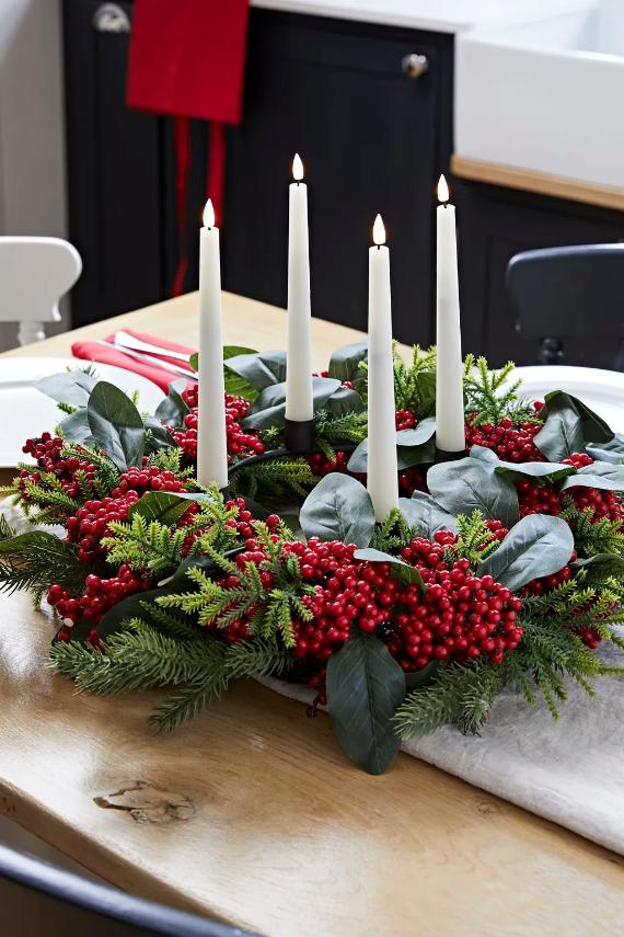 Festive wreath centerpiece with evergreen branches, red berries, eucalyptus, and four lit white candles, evoking a cozy, warm holiday ambiance.