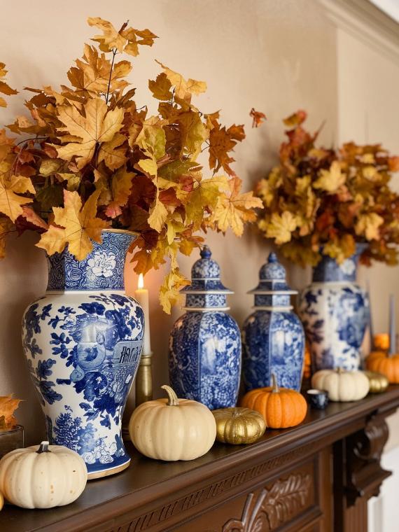 Blue and white vases with autumn leaves are displayed on a wooden mantel, surrounded by small orange, white, and gold pumpkins, creating a cozy blue fall decorations for mantel.