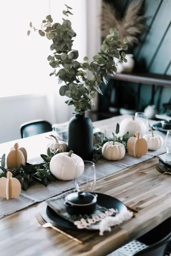 Elegant autumn-themed dining table with small white pumpkins, greenery, and a black vase with lush foliage. Greenery black plates, glasses, and gold utensils add a refined touch.
