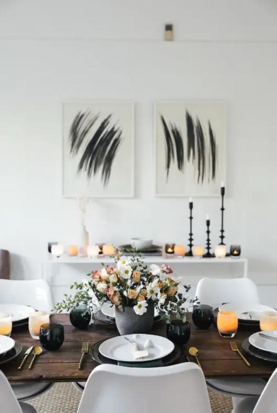 A modern dining room with a wooden table set with plates, gold utensils, black glasses, and orange candles, featuring a floral centerpiece and abstract artwork on the wall.