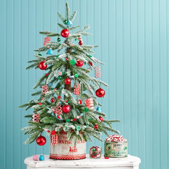 Small, decorated Christmas tree with red ornaments, candy-themed garlands, and snow-dusted branches on a white table. Blue paneled wall background.