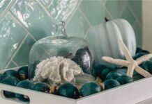 66 Inventive Seasonal Thanksgiving Décor Ideas With Blue A white tray holds teal ornaments, a starfish, a light blue pumpkin, and a glass dome with coral. Background features teal tiled walls, creating a coastal feel.