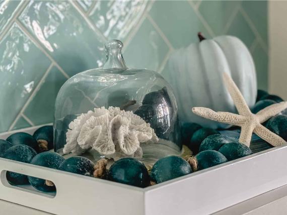 A decorative tray with coastal Thanksgiving decor with blue teal ornaments holds a glass dome, coral, a white pumpkin, and a starfish. Background has glossy aqua tiles, creating a coastal vibe.