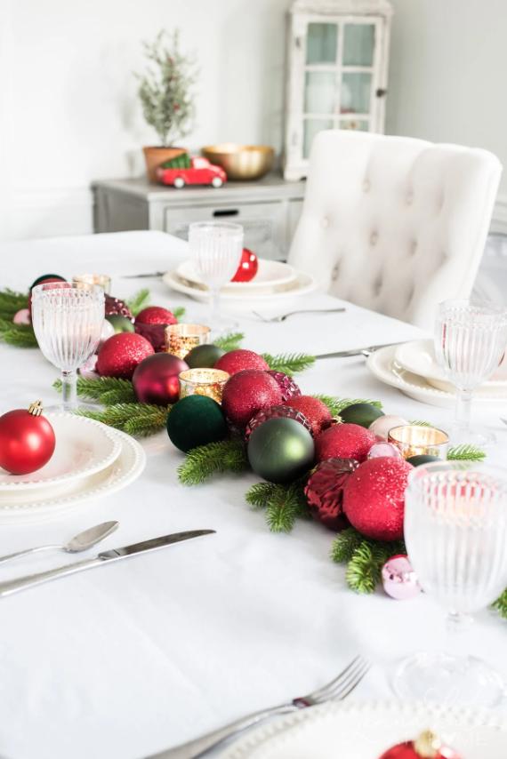 Elegant holiday table setting with a white cloth, red and green ornaments, pine branches centerpiece, crystal glasses, and white plates. Festive and inviting.