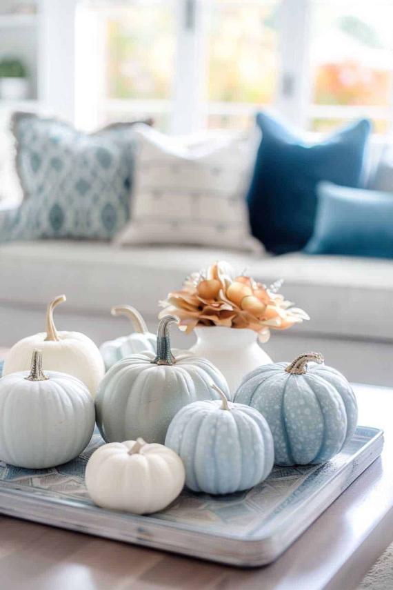 A decorative tray with white and pale blue pumpkins on a table. Blurred background with a cozy sofa and blue patterned cushions. Soft autumn vibe.