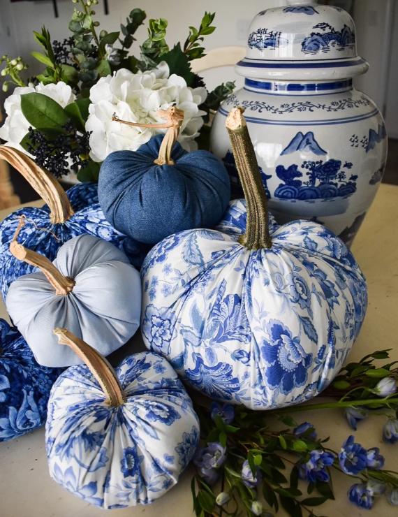 Blue and white fabric pumpkins with floral patterns are arranged near a matching ceramic vase and white flowers, creating an elegant autumnal setting.