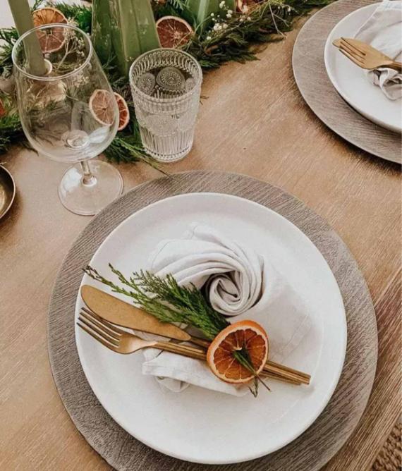 Elegant table setting with a white plate on a woven placemat, featuring a napkin, gold cutlery, greenery, and a dried orange slice. Nearby, a glass and greenery.