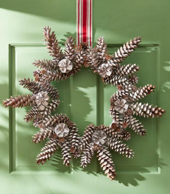 A festive wreath made of pine cones hangs on a green door, with a red and white striped ribbon. The scene conveys a rustic and cheerful holiday mood.
