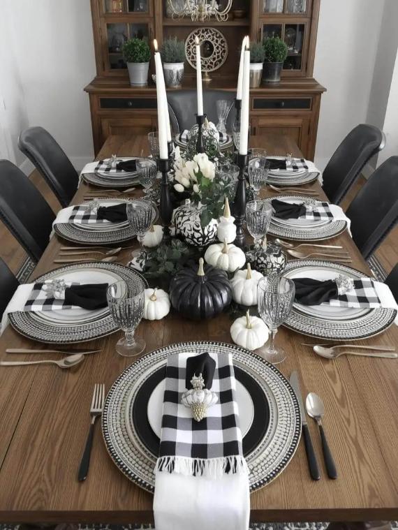 Dining table set with black and white theme, featuring layered plates, checkered napkins, pumpkins, and candles.