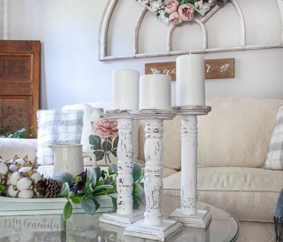 A cozy living room with a vintage chic vibe. White candles on rustic holders sit on a glass table. A couch with floral and plaid pillows is behind.