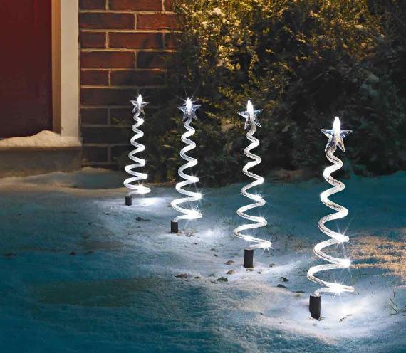 Snowy path illuminated by four glowing, spiral Christmas lights shaped like trees with star tops. Brick wall and bushes create a cozy, festive ambiance.