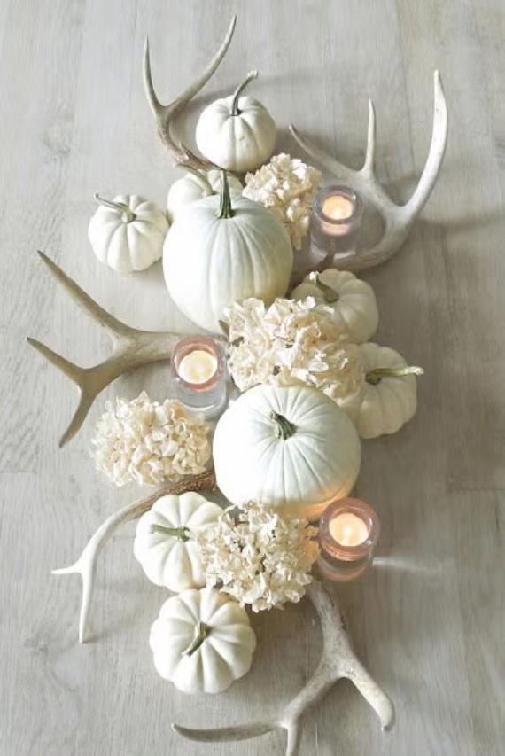 White pumpkins, soft beige hydrangeas, and antlers are arranged on a light wooden surface. Small candles add a warm, cozy glow.