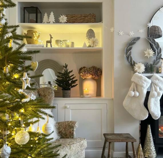 Cozy Christmas scene with a lit tree, white stockings by a wreath-adorned fireplace, and illuminated shelves displaying festive ornaments and decor. Warm, inviting ambiance.
