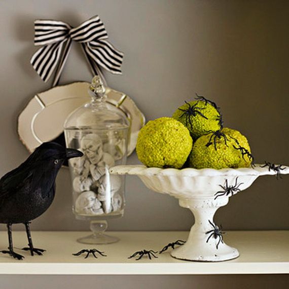A black bird perches on a shelf beside a bowl of green apples and a small spider.