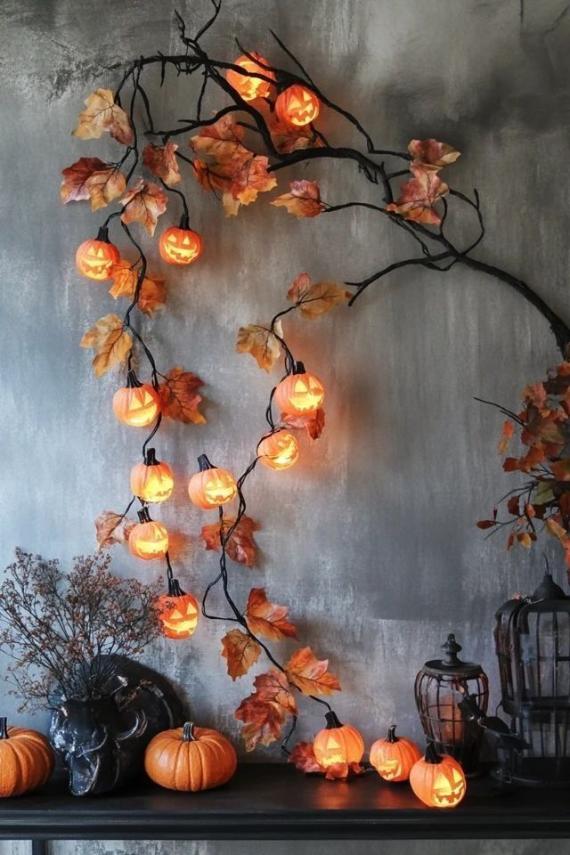 A textured gray wall featuring a large, dark branch arching across the top with glowing mini pumpkin string lights and autumn leaves cascading downward onto a table.