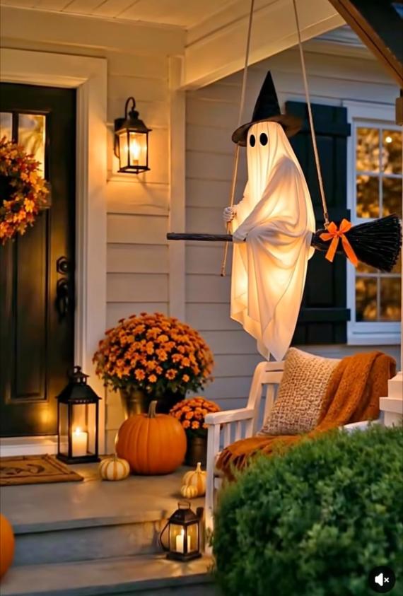 A cozy front porch decorated for Halloween featuring a glowing hanging sheet ghost wearing a witch hat and riding a broomstick, surrounded by pumpkins and orange mums.