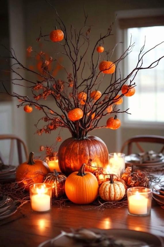  A wooden dining table styled with a large pumpkin centerpiece holding tall, bare branches adorned with hanging miniature orange pumpkins, surrounded by glowing candles.