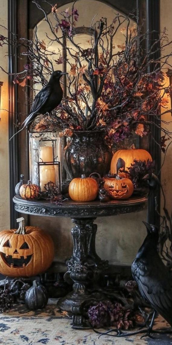 An ornate black pedestal table decorated for Halloween with a tall vase of dark, dried branches, realistic black crows, glowing glass lantern candles, and assorted carved orange pumpkins.