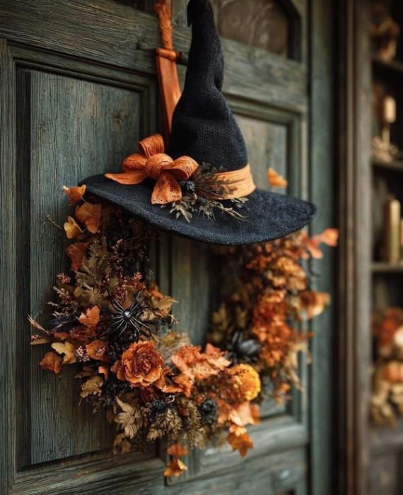 A rustic Halloween wreath made of dried orange leaves, dark pinecones, and black spiders, crowned with a large black fabric witch hat on a weathered green door.