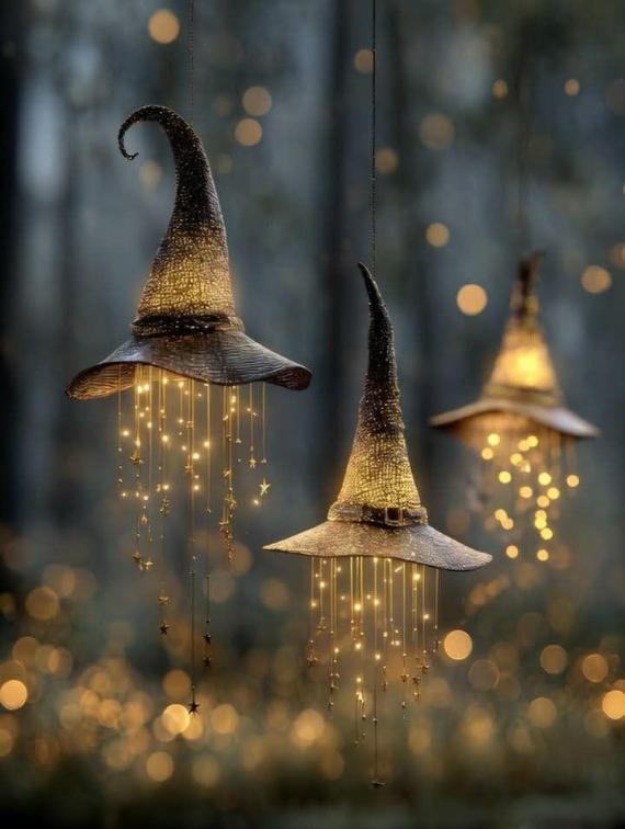 Three textured brown witch hats hanging in the air with glowing fairy lights and tiny metallic stars cascading from beneath the brims against a blurry, warm bokeh background.