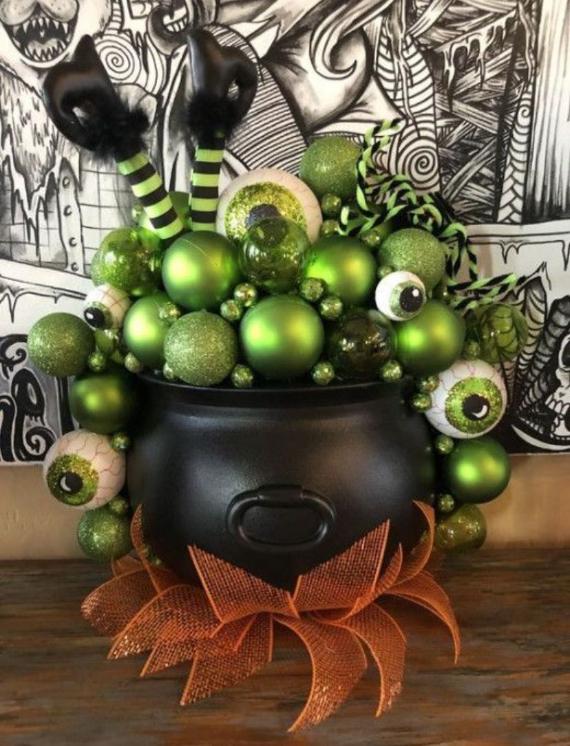 A matte black witch's cauldron centerpiece overflowing with green ornaments, large bloodshot eyeballs, and striped witch legs, sitting on an orange mesh ribbon base.