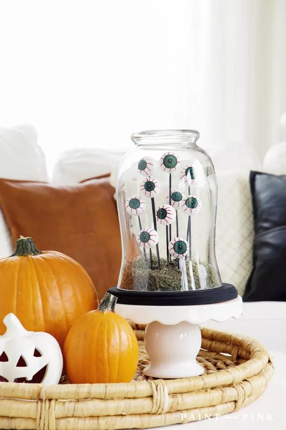 A glass dome decorated with Eyeballs containing colorful pumpkins alongside a rustic wicker basket on a wooden surface.