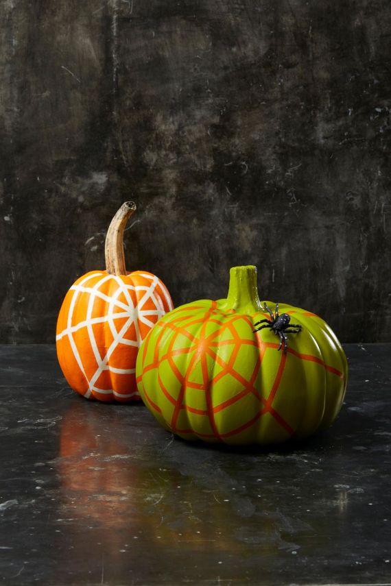 Two painted pumpkins with spiderweb designs, one orange with a white web and one green with an orange web and a faux black spider, against a dark background.