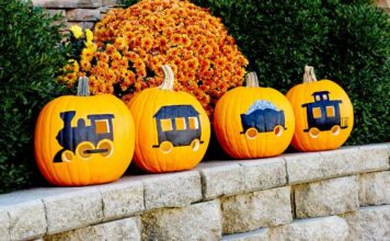 40 Eerily beautiful Halloween decoration Four orange pumpkins painted and carved to look like a train locomotive and cars, lined up on a stone retaining wall in front of an orange chrysanthemum bush.
