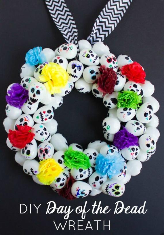 A handmade circular holiday door wreath made from dozens of small white painted sugar skulls, accented with vibrant crepe paper flowers and hanging from a black and white chevron ribbon.