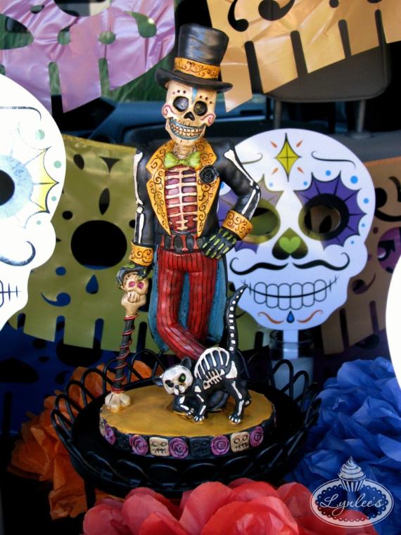 A hand-painted Day of the Dead skeleton figurine of a dapper Catrín man wearing a black top hat, a red striped suit, and a green bow tie, standing next to a skeleton cat on a pedestal display.