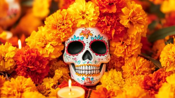 A hand-painted white sugar skull adorned with blue, pink, and orange floral patterns, nestled tightly inside a dense bed of vibrant orange and yellow marigold flowers with a lit candle in the foreground.
