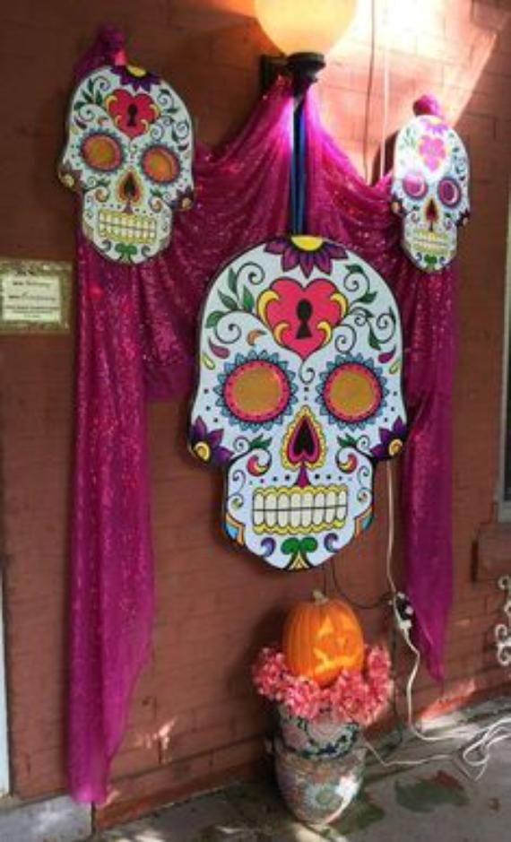 A large, colorful cut-out sugar skull wall hanging adorned with a heart lock design, framed by shimmering magenta fabric drapery and flanked by two matching smaller skulls on an outdoor brick wall.