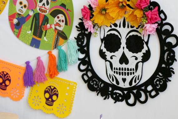  A black laser-cut circular wall wreath featuring a white sugar skull wearing a crown of yellow and pink fabric flowers, hanging alongside colorful paper banners and yarn tassels.
