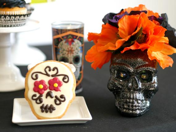  A cracked black mosaic skull vase topped with bright orange faux flowers, sitting on a black tablecloth next to an iced sugar skull cookie on a small square plate.