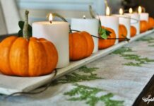 45 Fall, Halloween and Thanksgiving Crafts A row of small orange pumpkins and white candles on a wooden tray creates a cozy, autumnal centerpiece on a patterned table runner.