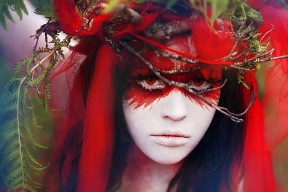 Modern Day witch of a woman with a painted white face and vivid red eye makeup wears a crown of branches, red cloth, and green leaves, evoking a mystical, ethereal mood.