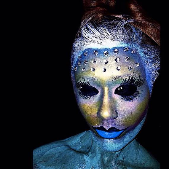 Mystical Glow Halloween Makeup of a person with intricate face paint featuring blue and gold hues, long eyelashes, and rhinestones on the forehead. The look is artistic and fantasy-themed.