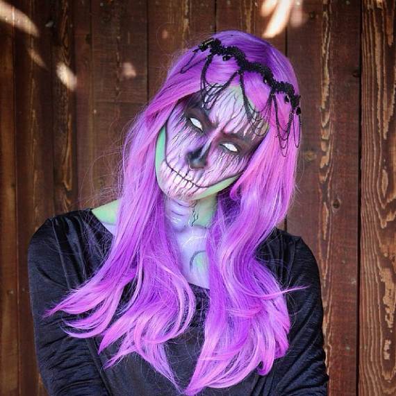 An Eerie Skull Queen Makeup  with purple hair and intricate skull face paint stares hauntingly. They wear a black headpiece and outfit, set against a wooden background.