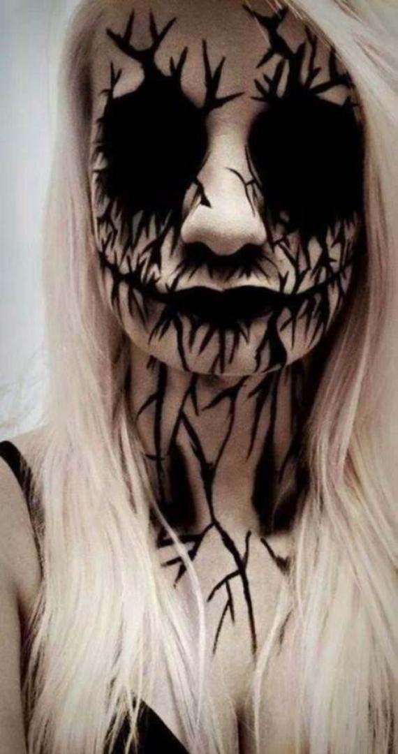 Close-up of a truly scary Halloween makeup idea with this chilling face artwith long blonde hair and dark, intricate face paint creating a haunting, cracked effect around eyes and mouth, extending to the neck. Eerie and dramatic.