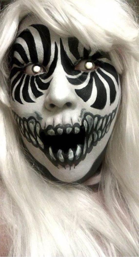Close-up of a Striped Demon Halloween Makeup person with intricate black and white face paint resembling a zebra pattern and skull, intense eyes, and a wide, toothy grin. They wear a white wig, conveying a haunting, eerie tone.
