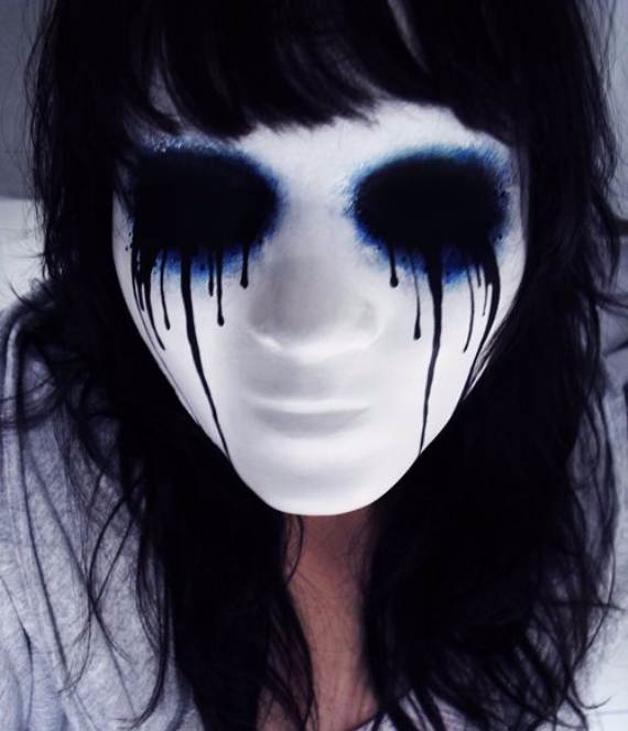 Person wearing a white mask with black, dripping eye makeup and blue accents, surrounded by dark hair. The Melting Eyes Makeup mood is eerie and mysterious.