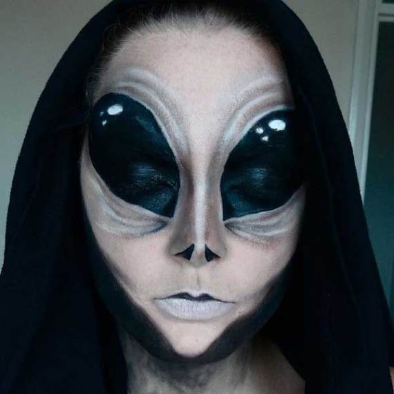 A person with Spooky Alien Halloween Makeup , featuring large, dark, reflective eyes and a pale face. They wear a black hood, creating a mysterious, eerie tone.