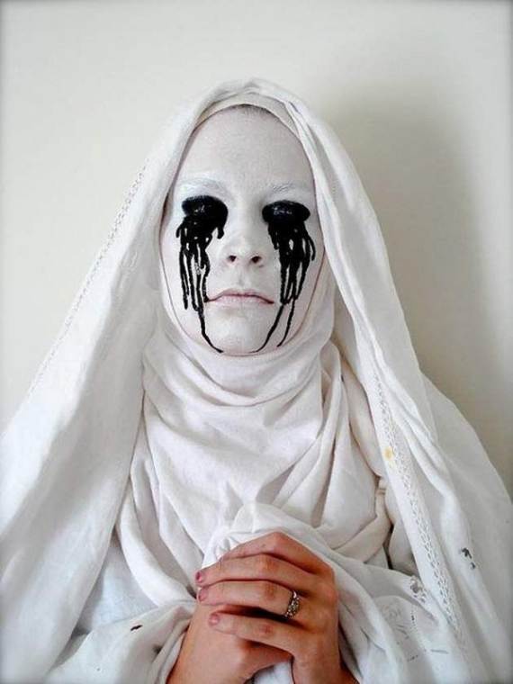 An easy scary Halloween makeup of a person with a white painted face and dramatic black tears wears a white robe and hood. Hands are folded in front, creating a haunting, somber tone.