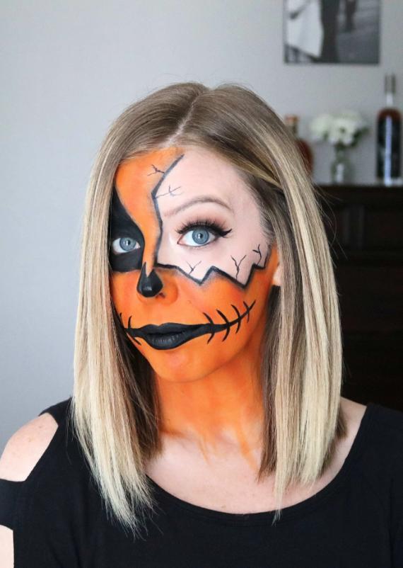 Woman with Cracked Pumpkin face paint, featuring one side with orange skin, black eye patch, and cracked details. Her expression is confident, wearing a black top.