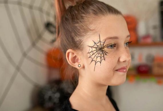 Young girl with a scary makeup ideas for Halloween. A spiderweb face paint by her eye, smiling softly. Background shows a playful Halloween theme with blurred decorations.