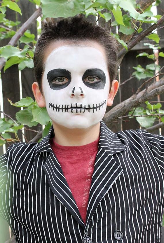A child with an Easy Jack Skellington Halloween Makeup of a skeleton face paint, featuring black eyes and a stitched mouth, wears a black and white striped jacket. They stand in front of a leafy background, conveying a playful Halloween vibe.