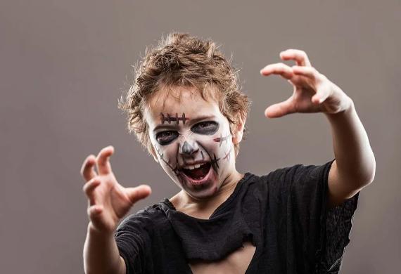 Child in aZombie makeup for kids ... torn black shirt with spooky face paint, resembling a skull and stitches, reaches forward playfully. The child appears excited and playful.