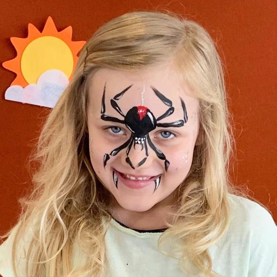 A child with Little Spider Face smiles, featuring a large spider face paint design with a red detail. The background has a paper sun and cloud on a red surface.